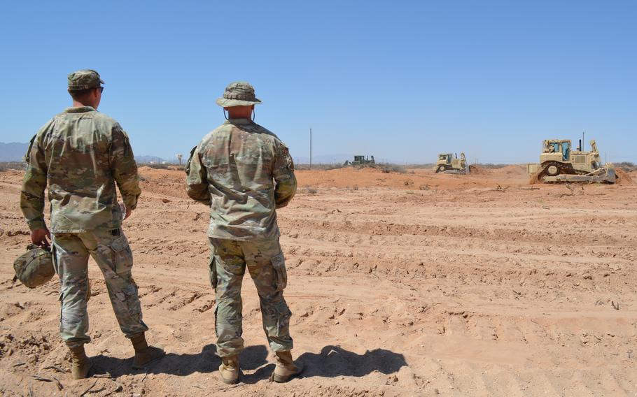 Two soldiers in camouflage look at a barren area of desert with bulldozers in the distance.