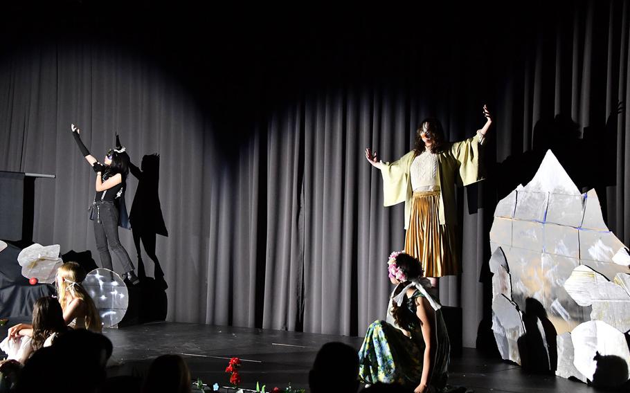 Students in costumes hold out their hands while performing against a black curtain on a stage.