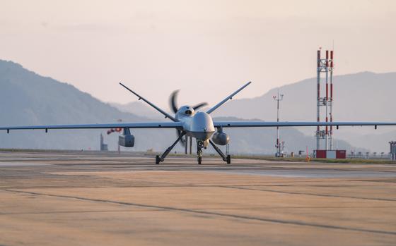 A military drone aircraft with long wings and a spinning propeller sits on a concrete runway at an airbase, with mountains and communication towers visible in the background under an overcast sky.