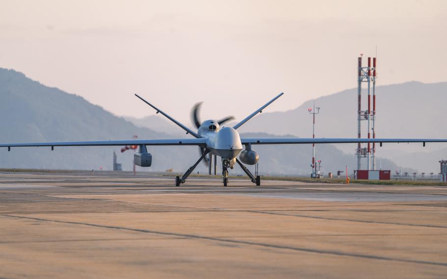 A military drone aircraft with long wings and a spinning propeller sits on a concrete runway at an airbase, with mountains and communication towers visible in the background under an overcast sky.