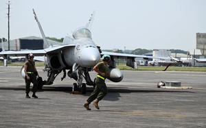Ground crew members work around a parked F/A-18 fighter jet on the runway, one crew member jogging while another stands by the aircraft.