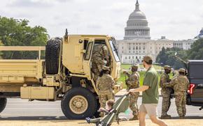 A man pushing a baby stroller walks on a sidewalk past a military vehicle with troops in camouflage uniforms standing in front, with the U.S. Caiptol building in the background.