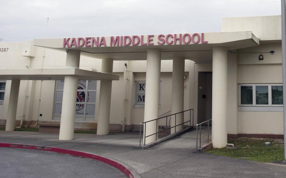 The front entrance of a school is seen, with lettering reading “Kadena Middle School” on an overhang above three support columns.