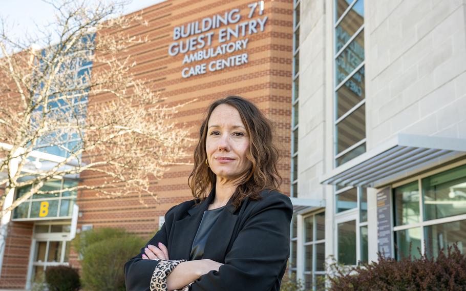 A woman in a dark blazer poses with her arms crossed in front of the entrance to a hospital.