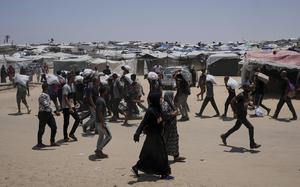 People carry bags of supplies with a makeshift village in the background. 