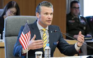 Pete Hegseth gestures with his arms spread while sitting at a table in front of a small U.S. flag and speaking into a microphone.