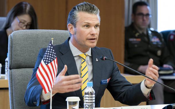 Pete Hegseth gestures with his arms spread while sitting at a table in front of a small U.S. flag and speaking into a microphone.