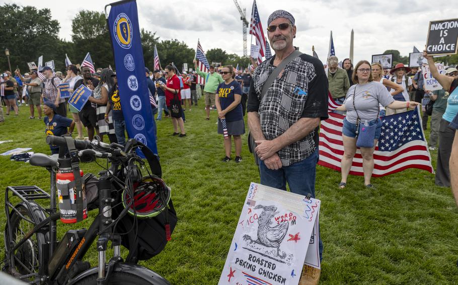 A man wearing sunglasses and a bandana stands in a grassy field with a protest sign leaning against his legs and other protesters behind him.