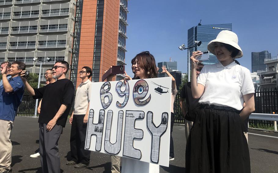 People gather outdoors in an urban setting, with one person prominently holding a large white sign that reads “BYE HUEY” with what appears to be a small helicopter silhouette. 