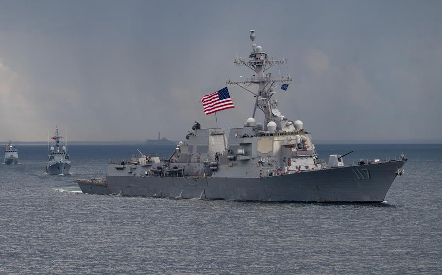 A naval destroyer flying a U.S. flag sails in open ocean with other warships in a line behind it and the horizon in the background.