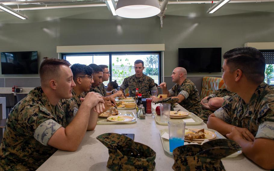 Several U.S. Marines, dressed in standard camouflage uniforms, are seated around a table in a dining facility, engaging in discussion while eating their meals. 