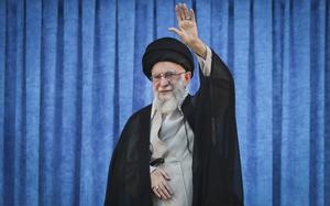 An Iranian man wearing a black robe, black turban and glasses waves with a raised hand as he stands in front of a blue curtain.