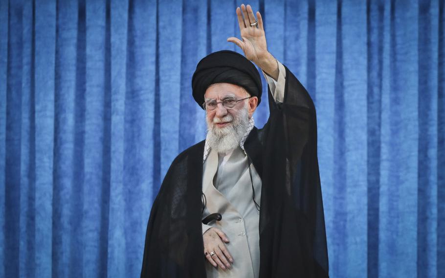 An Iranian man wearing a black robe, black turban and glasses waves with a raised hand as he stands in front of a blue curtain.