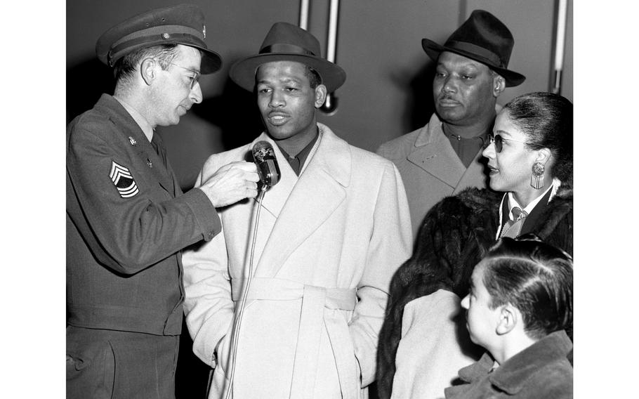 A black and white photo of a man speaking into a microphone as others look on.