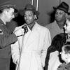 A black and white photo of a man speaking into a microphone as others look on.