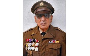 An elderly man poses in front of a white wall while wearing a brown military dress uniform, hat and sunglasses.