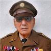 An elderly man poses in front of a white wall while wearing a brown military dress uniform, hat and sunglasses.