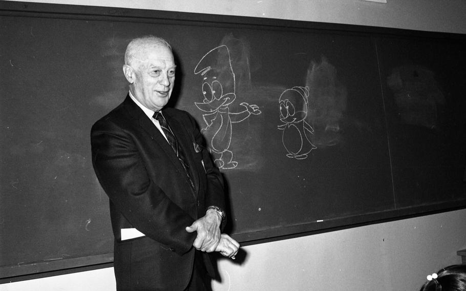 A black and white photo of a man in a suit standing in front of a blackboard with cartoon birds drawn on it.