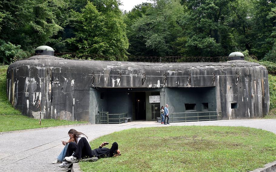 Exterior view of a concrete World War II bunker with two observation domes, surrounded by green grass and trees, with visitors walking near the entrance.