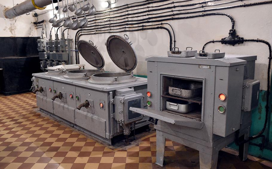 Military kitchen equipment including large cooking pots, ovens and preparation surfaces with control panels and indicator lights, preserved in an underground facility.