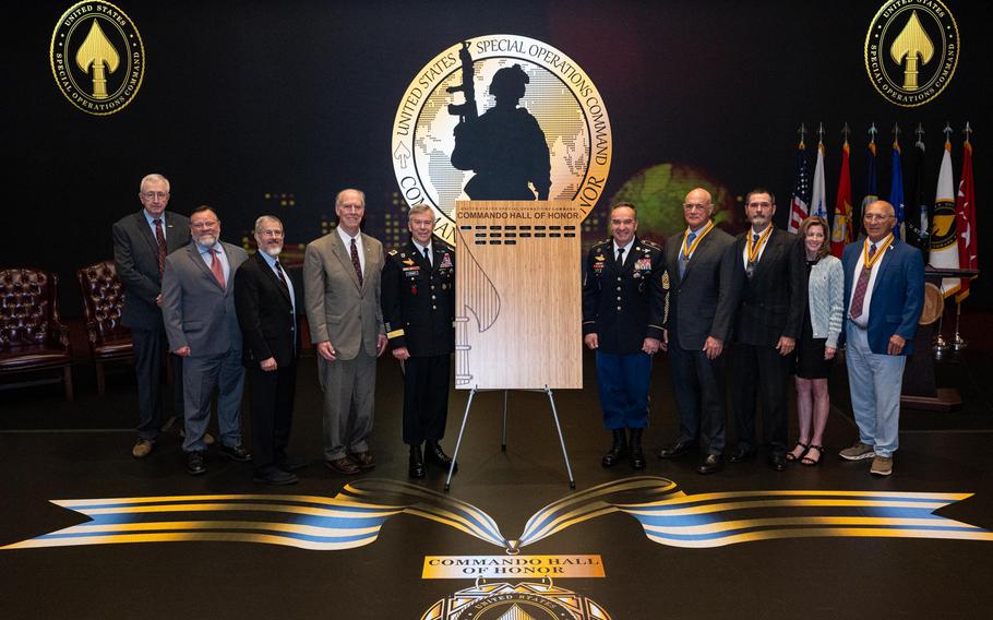A line of people in military uniforms or suits stand on either side of a wooden board bearing names.