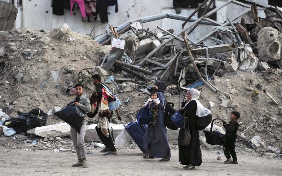 Palestinian people carrying their few belongings in sacks while walking past destroyed buildings.