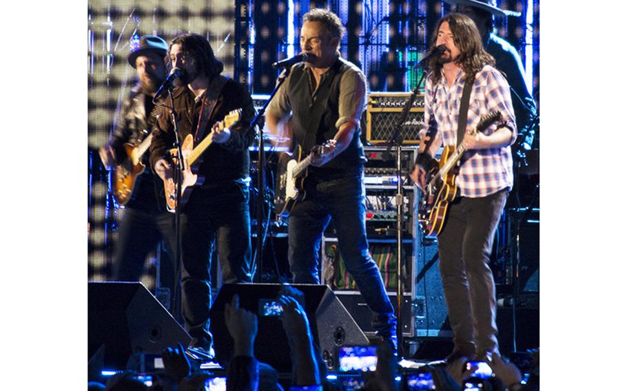 Several people perform on stage, four in the front with guitars and one in the back who is mostly obscured.