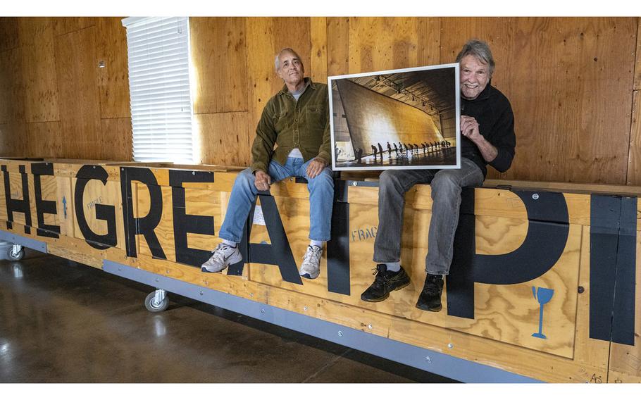 Photographers Clayton Spada, left, and Jacques Garnier hold a photograph of “The Great Picture,” the world’s largest photograph as they sit on a 46-foot-long box that holds the photograph at the Great Park Artist Studios in Irvine on Feb. 17, 2023. The image shows a portion of the former Marine Corps Air Station El Toro. 