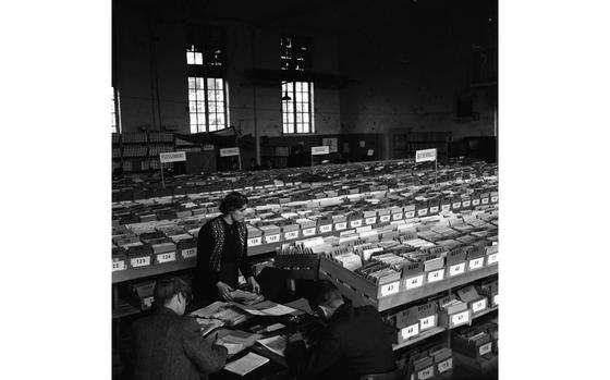 Esslingen, West Germany, Sep. 27, 1952: Clerks and investigators search through row upon row upon row of records in the massive factory-floor-turned records room of the International Tracing Service's offices in Esslingen, in Baden-Wuerttemberg. Signs marking the records from the various Nazi concentration camps can be seen in the back. 
Searching through the Child Search master index of some 204,000 cards which include 70,750 birth certificates the investigators search for non-German children separated from their families during the war in an effort to reunite them with their families.

Read Stars and Stripes' article on the post-war efforts of reunification by the ITS here. 

The International Tracing Service morphed into the Arolsen Archives and its staff continues the work of their 1950s colleagues began. “The Arolsen Archives are committed to preserving their unique collection of documents on Nazi persecution and to making them accessible worldwide. We search for the traces of victims and survivors of the National Socialist terror regime and, even today, we are still helping to reunite families torn apart by the Holocaust, the persecution of minorities and forced labor.”
For more information about the organization, its archives, and work, visit their website.[https://arolsen-archives.org/]
