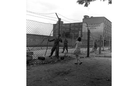 A West Berlin girl looks on as young, uniformed East Germans roll out barbed wire
