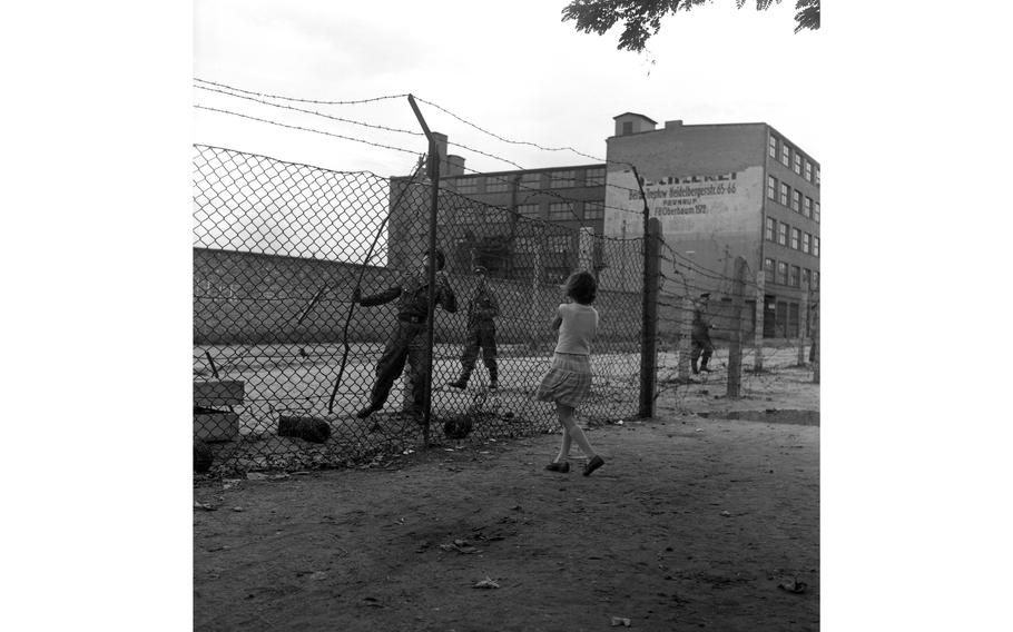 A West Berlin girl looks on as young, uniformed East Germans roll out barbed wire.