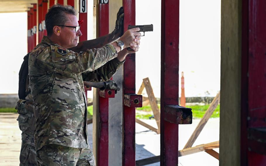 A military officer in camouflage uniform and dark-rimmed glasses points a handgun between red posts set up on a shooting range.