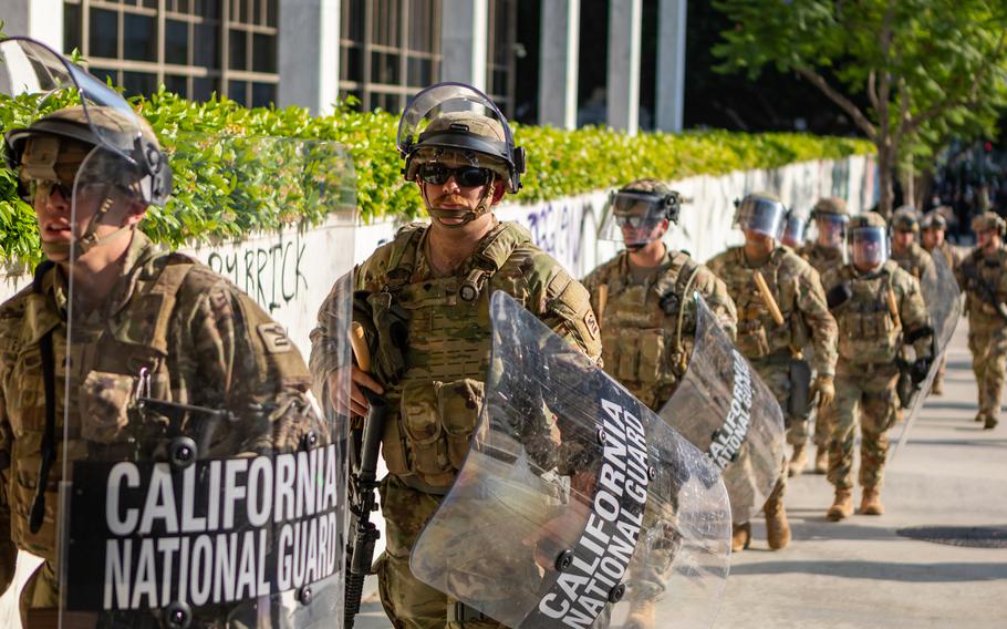 National Guard members carrying shields walk past a wall covered in graffiti. 