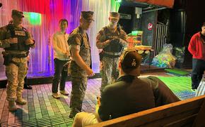 Military police and  Col. David Banning interact with service members on a  street at night, near a colorful curtain backdrop.