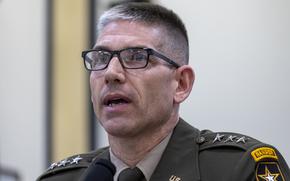 Close-up view of an Army officer wearing a dress uniform and black glasses speaking into a microphone at a congressional hearing.