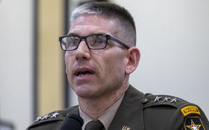 Close-up view of an Army officer wearing a dress uniform and black glasses speaking into a microphone at a congressional hearing.