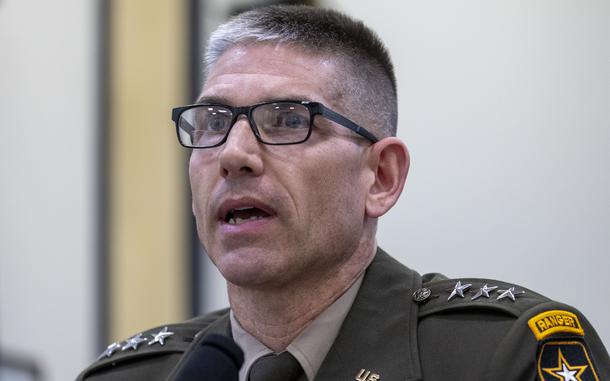 Close-up view of an Army officer wearing a dress uniform and black glasses speaking into a microphone at a congressional hearing.