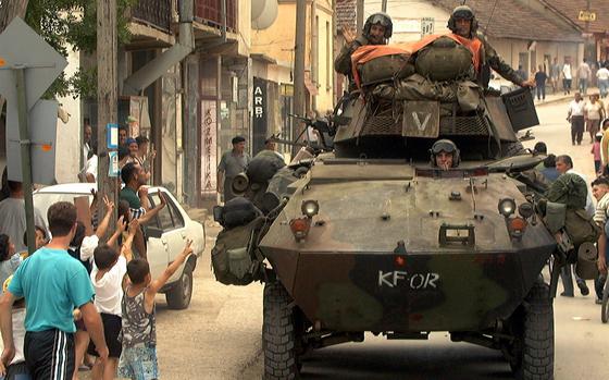 Children wave to a tank rolling down a street with military personnel visible atop it.