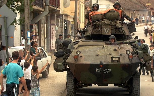 Children wave to a tank rolling down a street with military personnel visible atop it.