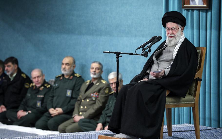 Ali Khamenei sits in a chair in front of a microphone, with military commanders seated behind him and to his left.