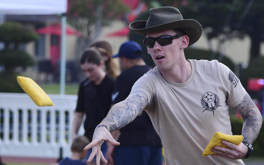 A man in a cowboy hat and dark sunglasses tosses a yellow bean bag with his right hand while holding another yellow bean bag in his left hand at an outdoor festival.
