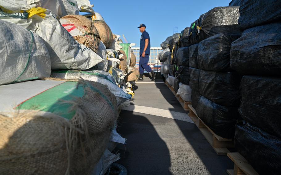 A Coast Guard crew member walks in the background between the bags of drugs.