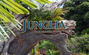 The entrance sign to “Junglia Okinawa” in blue and gold lettering, positioned on a natural rock archway surrounded by tropical vegetation, including palm fronds and dense green foliage.