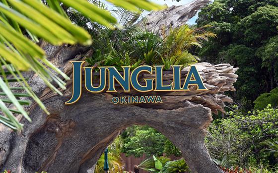 The entrance sign to “Junglia Okinawa” in blue and gold lettering, positioned on a natural rock archway surrounded by tropical vegetation, including palm fronds and dense green foliage.