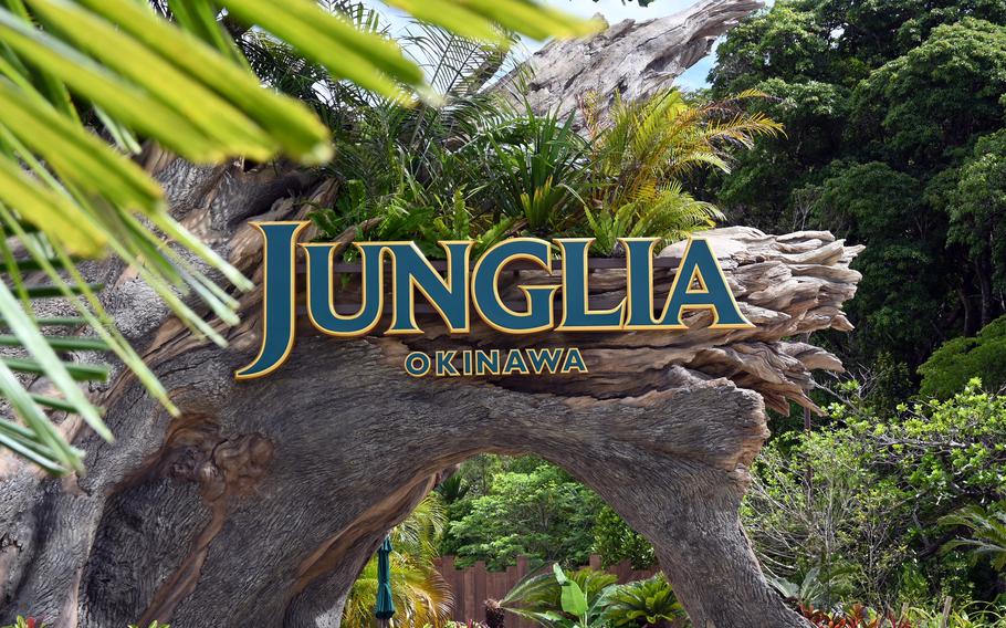 The entrance sign to “Junglia Okinawa” in blue and gold lettering, positioned on a natural rock archway surrounded by tropical vegetation, including palm fronds and dense green foliage.