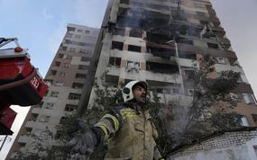 A firefighter at the base of a building gestures; the building towering above him is plainly damaged.