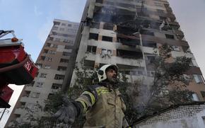 A firefighter at the base of a building gestures; the building towering above him is plainly damaged.