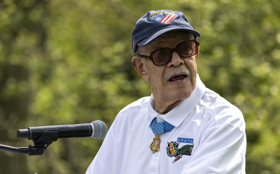 An elderly man in a white shirt, blue hat and sunglasses stands at a microphone and speaks during a ceremony.