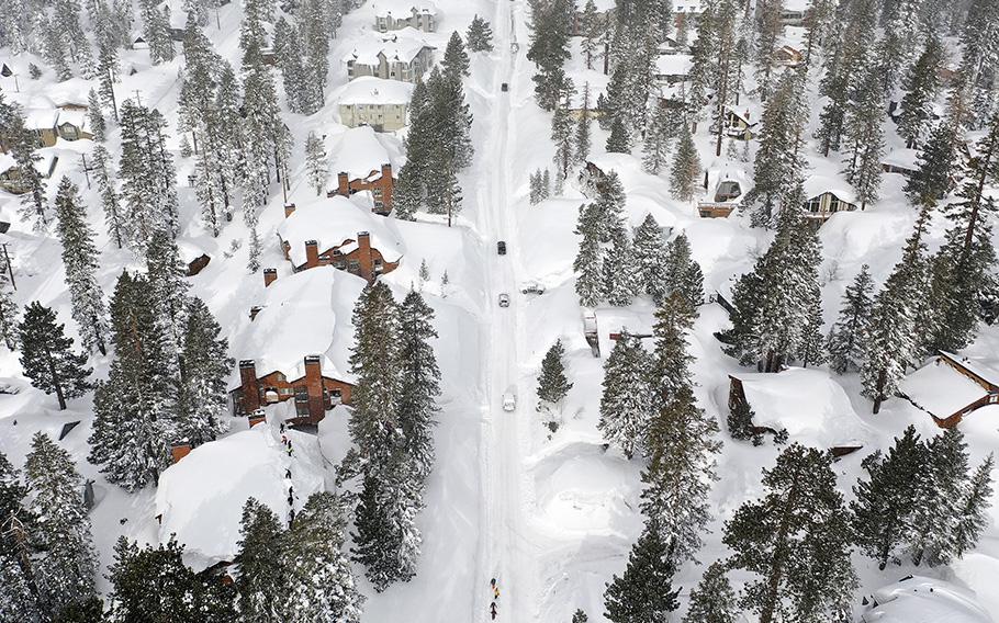 Snowboarders walk and cars drive in the Sierra Nevada mountains, near snowbanks piled up from new and past storms, after yet another storm system brought heavy snowfall further raising the snowpack on March 29, 2023, in Mammoth Lakes, California. 