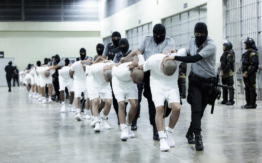 Masked guards hold prisoners’ hands behind their backs and their heads down as they walk them in a line.